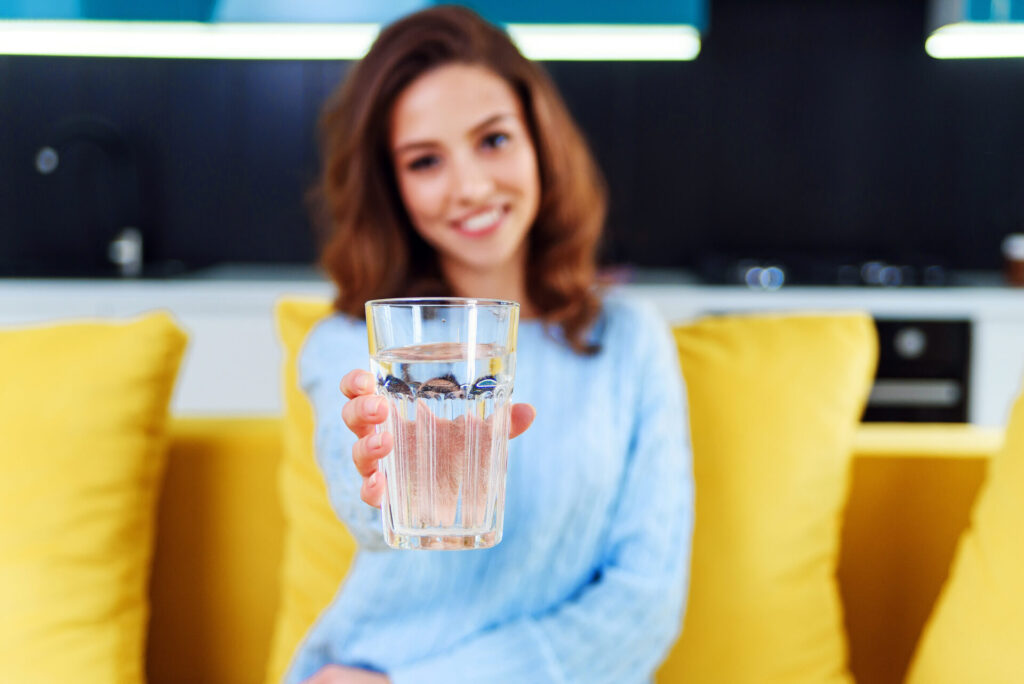 Eine junge, glückliche Frau in einem blauen Pullover sitzt auf einer gelben Couch und bietet ein Glas kristallklares Wasser an.