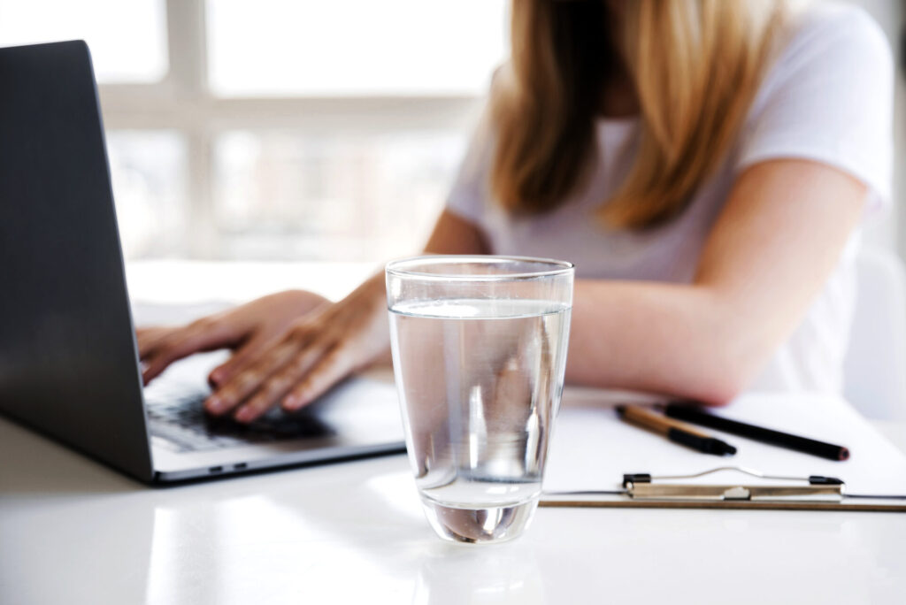 Ein Glas mit klarem, gefiltertem Wasser steht auf einem Schreibtisch neben einer Person, die an einem Laptop arbeitet, um hydriert und konzentriert zu bleiben.