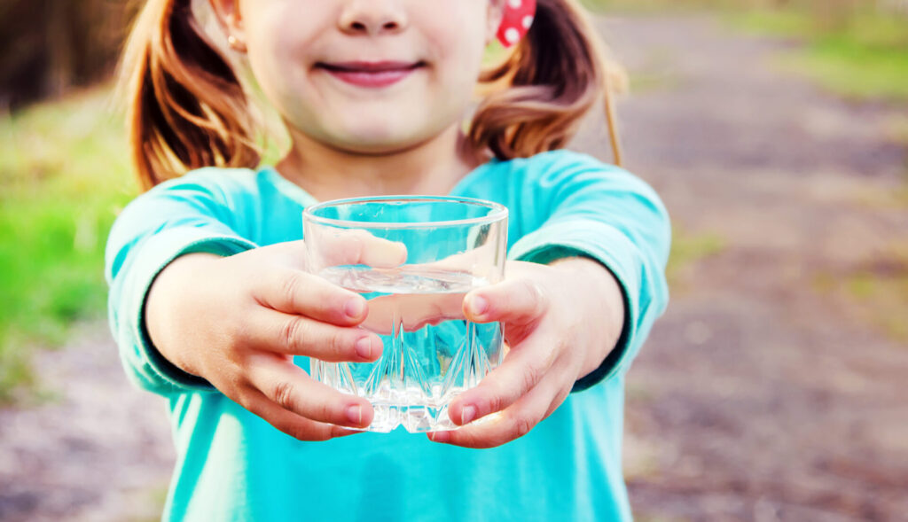 Ein lächelndes Mädchen in einem türkisen Oberteil reicht ein Glas mit frischem, klarem Wasser nach vorn.
