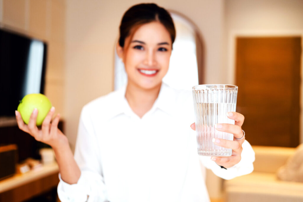 Eine lächelnde Frau in einem weißen Hemd hält ein Glas klares, frisches Wasser und einen grünen Apfel in die Kamera, um einen gesunden Start in den Tag zu symbolisieren.