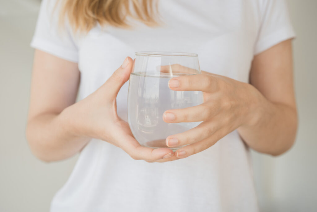 Eine Frau in einem weißen T-Shirt hält ein Glas mit klarem, reinem Wasser in beiden Händen.