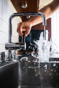 Frisches Wasser spritzt aus einem Glas, das unter einem laufenden Küchen-Wasserhahn überläuft.
