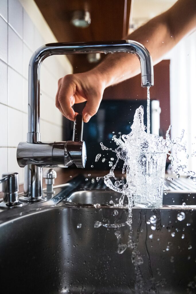 Frisches Wasser spritzt aus einem Glas, das unter einem laufenden Küchen-Wasserhahn überläuft.
