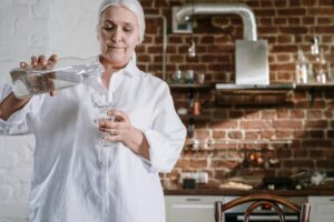 Eine Seniorin mit weißem Haar, bekleidet mit einem weißen Hemd, schenkt sich in einer Küche mit Backsteinwand Wasser aus einer Glaskaraffe in ein Glas ein.