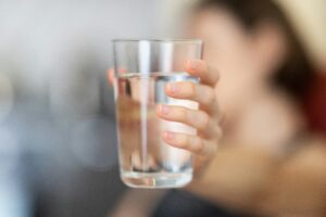 Eine Hand hält ein Glas mit klarem, reinem Wasser in die Kamera, der Hintergrund ist unscharf.