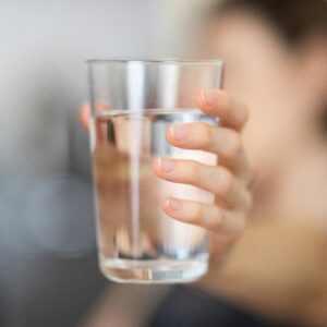 Eine Hand hält ein Glas mit klarem, reinem Wasser in die Kamera, der Hintergrund ist unscharf.