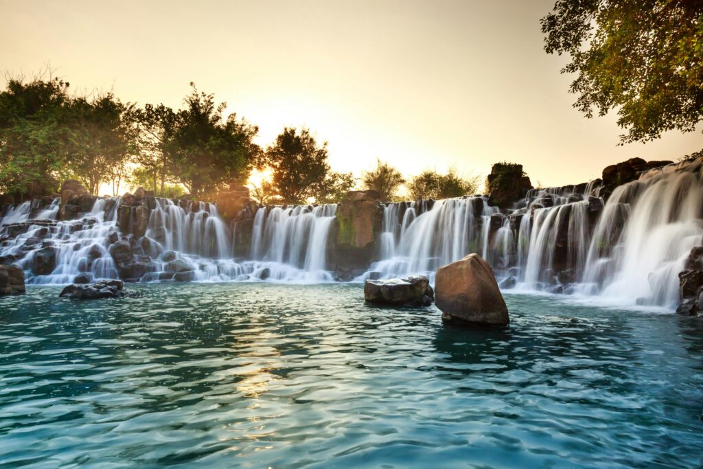 Ein breiter Wasserfall fließt in Kaskaden über Felsen in einen klaren See bei Sonnenuntergang.